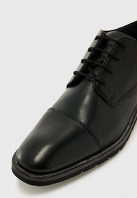 Zapato de vestir de cuero negro con acabado pulido, puntera redondeada y cordones estándar. Suela texturizada y detalles de costura sutiles.