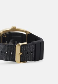Correa de reloj de tela negra con acabado texturizado, hebilla en tono dorado y caja de acero inoxidable. Presenta un diseño minimalista y agujeros ajustables.
