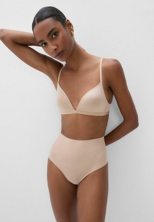 Kvinde med mørk hud og håret sat tilbage bærer beige bh og trusser med høj talje, poserer mod en hvid væg med den ene arm hævet.