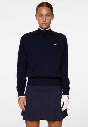 Marineblauer Pullover mit einem Viertelreißverschluss, gerippten Bündchen und Kragen. Kombiniert mit einem passenden Faltenrock. Das Modell trägt einen weißen Golfschuh.