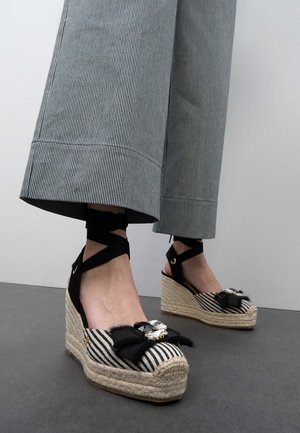 Pies que llevan alpargatas cuña a rayas con lazadas alrededor del tobillo y lazos decorativos, combinadas con pantalones anchos de rayas grises.