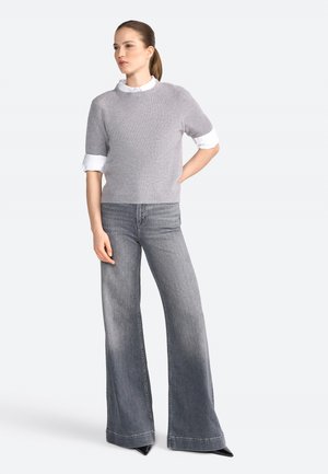 Maglione grigio a maniche corte a maglia indossato sopra una camicia bianca con colletto, abbinato a jeans larghi grigio chiaro e décolleté nere a punta.
