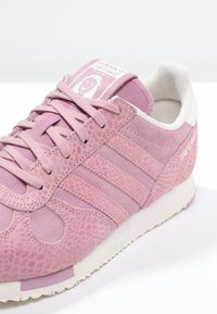 Rosa sneakers i mocka med en texturerad yta, med tre ränder på sidan, vit sula och en märkestungetikett på tungan.