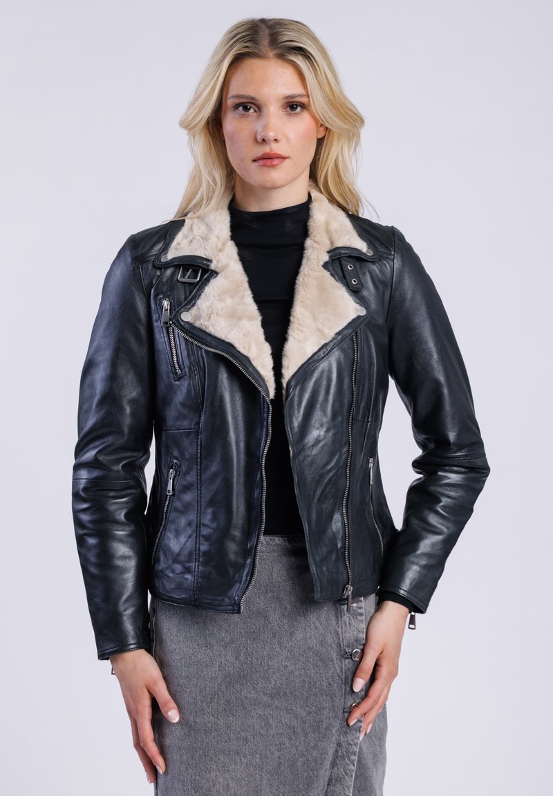 Mladá žena se světlými vlasy, která nosí černou koženou bundu s béžovým kožíškem na límci, černý top a šedou sukni, na jednobarevném světlém pozadí.