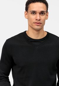 Zwarte gebreide trui met een v-hals ontwerp. Beschikt over lange mouwen en een soepele textuur. Gedragen over een zwarte ronde hals shirt.