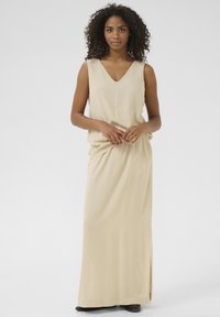 Robe maxi beige en tissu doux, avec un décolleté en V profond, sans manches et une fente sur le côté pour faciliter le mouvement. Texture lisse.