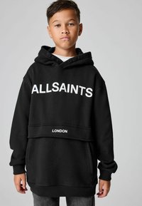 Chico vestido con una sudadera negra de "ALLSAINTS" con el texto "LONDRES", de pie frente a un fondo gris liso, mirando hacia adelante.