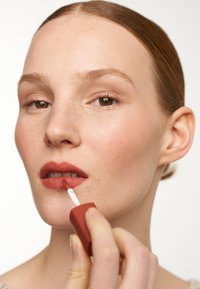 Matte liquid lip color in a rounded, terracotta-colored applicator, applied to lips. Smooth texture with a subtle sheen, hand visible.