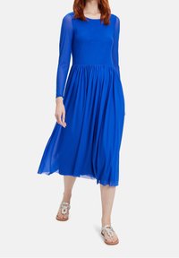 Blå klänning i medellängd med långa genomskinliga ärmar, en åtsittande överdel och en flödande veckad kjol. Modellen bär ljusa sandaler.