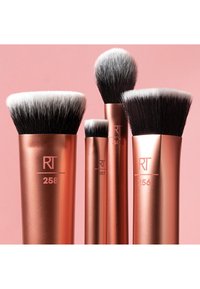 Quatre pinceaux de maquillage métalliques couleur or rose avec des poils denses noirs et blancs sur un fond rose, étiquetés RT avec des numéros.