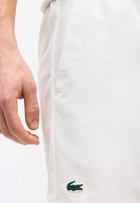 Κοντινό πλάνο ενός χεριού δίπλα σε λευκά σορτς με μικρό πράσινο λογότυπο κροκοδείλου κοντά στο τελείωμα.