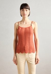 Débardeur couleur rouille à ourlet en dents de scie, motif zigzag texturé et bretelles métalliques. Associé à un pantalon crème.