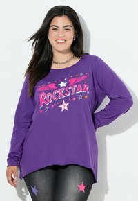 Smilende kvinde iført en lilla langærmet skjorte med "Rockstar" tekst og stjerner, parret med sorte leggings med lilla og pink stjerner.