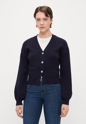Cardigan bleumarin cu decolteu în V, mâneci lungi tip puff și cinci închideri cu butoane metalice. Textură moale, talie ajustată și manșete ribbate.