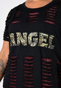 Černé tričko s krátkým rukávem, velkými výřezy a grafikou „ANDĚL“ s leopardím vzorem. Má kulatý výstřih a volný střih.