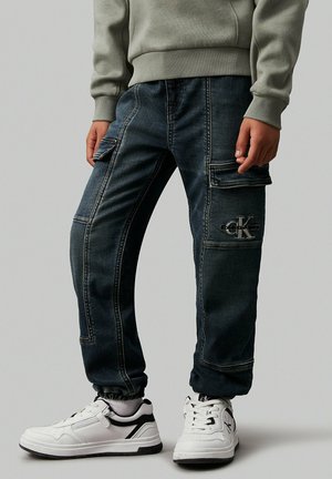 Osoba nosząca ciemnoniebieskie dżinsowe spodnie typu cargo z logo Calvin Klein i białe trampki na jednolitym szarym tle.