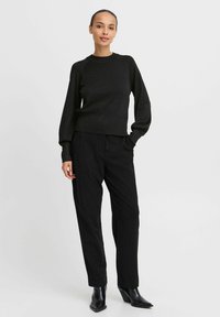 Schwarzer Strickpullover mit langen Ärmeln und rundem Halsausschnitt, kombiniert mit schwarzen, maßgeschneiderten Hosen mit kleinen gepunkteten Mustern und schwarzen Stiefeln mit Absatz.