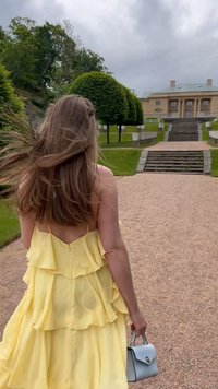 Gul, lagdelt volangkjole med lav rygg, kombinert med en liten lyseblå veske, i en anlagt hage med trapper og grøntområder.