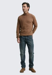 Brauner Rollkragenpullover aus weichem Strickstoff, kombiniert mit blauen Jeans. Das Modell trägt beigefarbene Wildleder-Stiefeletten. Einfaches, lässiges Design.