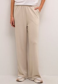 Pantalon beige à jambes larges avec une ceinture élastique, des poches latérales et une texture lisse et douce. Associé à des baskets blanches et beiges.