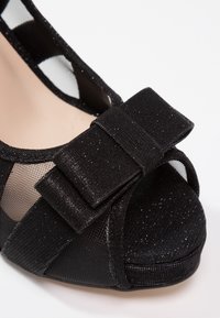 Chaussure noire à bout ouvert en maille avec un grand nœud en tissu et des détails subtils de paillettes sur un fond blanc.
