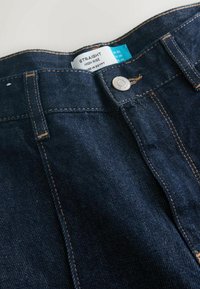 Dunkelblaue, gerade geschnittene High-Rise-Jeans mit goldenen Nähten, einem Metallknopf und einem sichtbaren Pflegeetikett am Bund. Der Stoff hat eine glatte Textur.