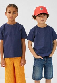 Des t-shirts en coton bleu marine à manches courtes, associés à un pantalon orange texturé et à un short en jean ; une casquette rouge présentée sur un enfant.