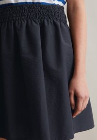 Zwarte rok met een smocktailleband, zacht vallende stof en een iets gestructureerd oppervlak, gedeeltelijk zichtbaar hand die de rok vasthoudt.