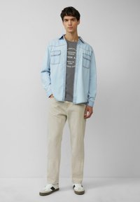 Lichtblauwe denimshirt, grijs graphic t-shirt, beige broek en witte sneakers met zwarte accenten. Twee borstzakken op het shirt.