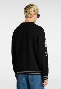 Suéter negro de punto con cuello redondo, que presenta acentos bordados en blanco en las mangas y una cinturilla a rayas. Combinado con jeans azules.