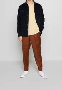 Zwart ribfluweel knoopshirt, lichtgele t-shirt, bruine geplooide broek en witte sneakers. Eenvoudig ontwerp met zakelijke zakken aan de voorkant.