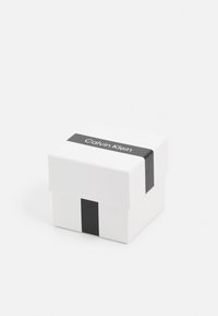 Hvit firkantet boks med en svart stripe rundt midten, som har merkevarenavnet "Calvin Klein" i hvit tekst på stripen.