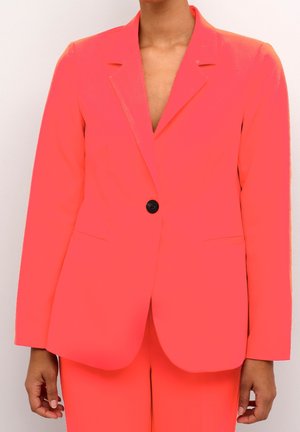 Vrouw die een fel koraalkleurig blazer draagt met één zwarte knoop en bijpassende broek, afgebeeld van nek tot heupen tegen een effen achtergrond.
