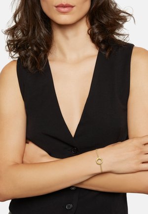 Gouden armband met een hexagonaal sieraad aan een delicaate ketting, gedragen op een arm gekruist over een zwarte mouwloze top.