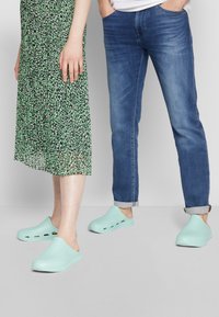 Tamancos azul claro com buracos de ventilação laterais e solas texturizadas, usados por duas pessoas ao lado de um vestido verde estampado e jeans azuis.