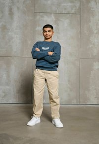 Szary sweter z napisem "ENGLEWOOD", beżowe chinosy i białe sneakersy. Pozycja stojąca w przeciwieństwie do gładkiej, teksturowanej szarej ściany.