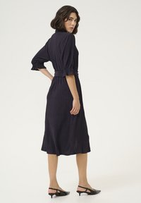Mörklila klänning med texturerad tyg, trekvartslånga ärmar med volangkragar, bälte i midjan och längd till mitten av vaden, parat med svarta slingback-skor.