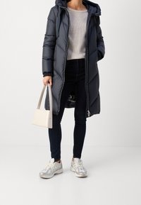 Abrigo acolchado en azul marino con capucha, cierre de cremallera y patrón acolchado. Llevado con un suéter de canalé, jeans oscuros y zapatillas blancas. Lleva un bolso beige.