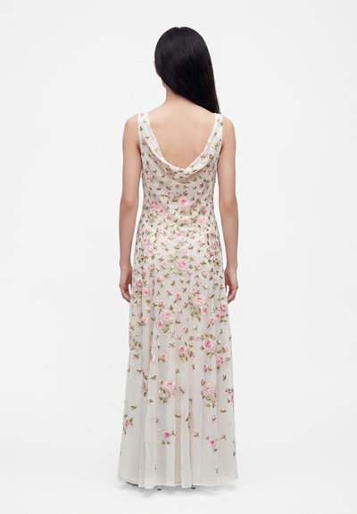 Femme aux longs cheveux foncés portant une robe blanche longue sans manches avec une broderie florale rose, vue de dos sur un fond uni.