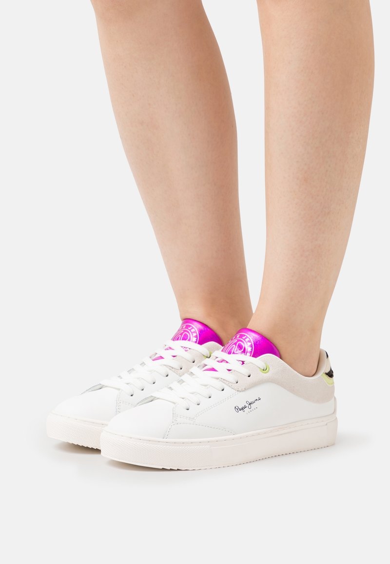 Bílé nízké tenisky s metalickým fialovým jazykem, bílými tkaničkami a nenápadným logem značky nošené na nahých nohách před jednobarevným pozadím.