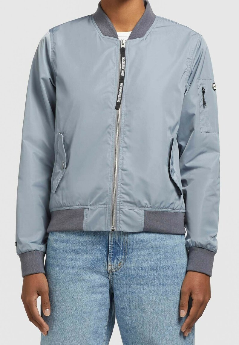 Veste bomber gris clair avec poignets, col et ceinture côtelés, fermeture éclair sur le devant, deux poches avant et une poche zippée sur la manche gauche.