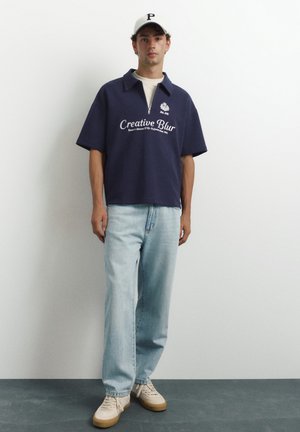 Jeune homme portant un polo zippé bleu marine à manches courtes avec un texte, un jean bleu clair, des baskets beiges et une casquette de baseball blanche, debout contre un mur blanc.