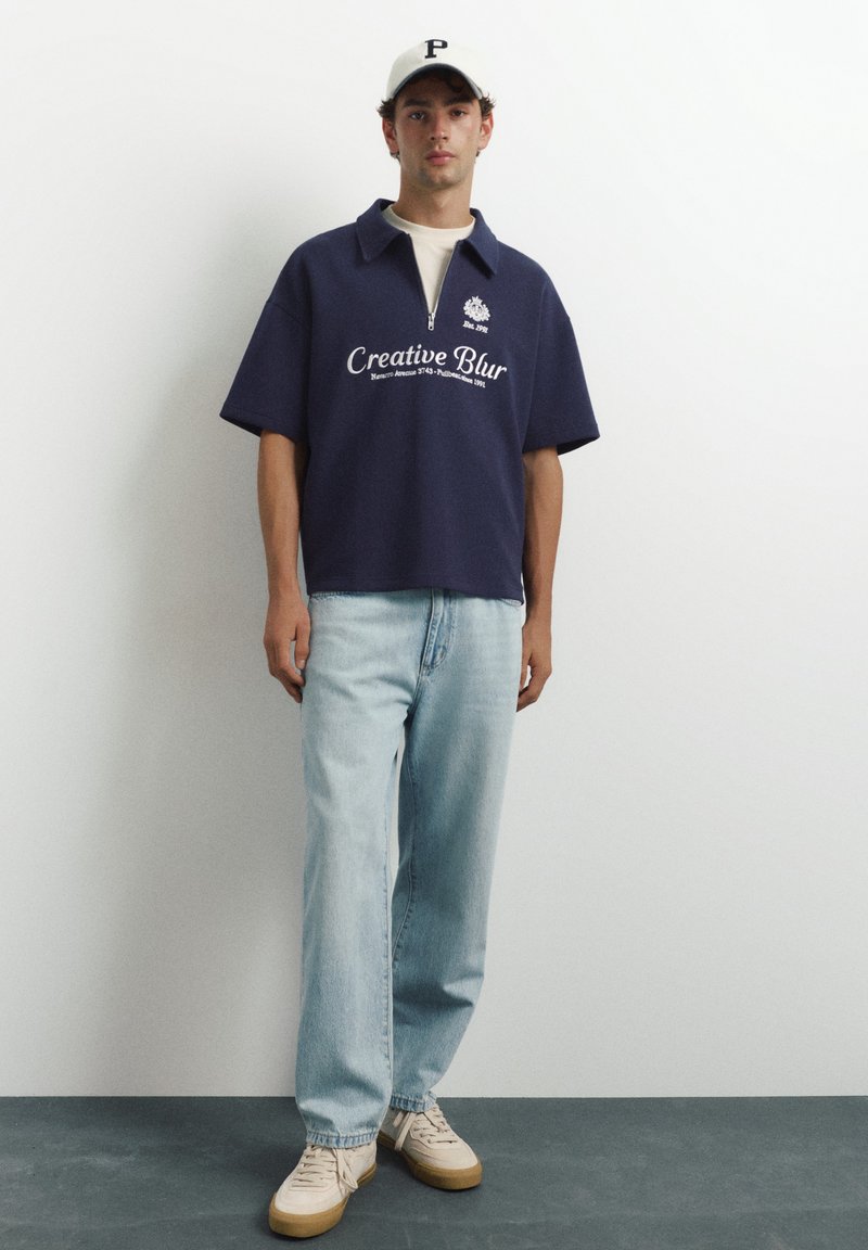 Jeune homme portant un polo zippé bleu marine à manches courtes avec un texte, un jean bleu clair, des baskets beiges et une casquette de baseball blanche, debout contre un mur blanc.