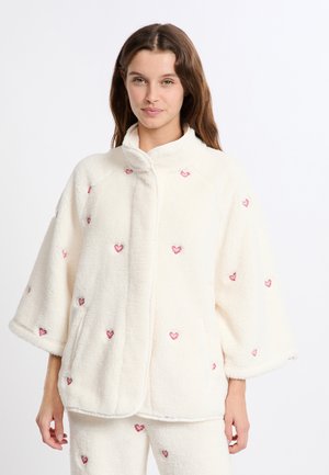 Chaqueta de forro polar crema con un ajuste grande y holgado y mangas de media longitud. Presenta patrones de corazones rojos y bolsillos laterales, con un diseño de cuello alto.