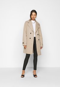 Casaco bege de dupla face com botões pretos, lapelas largas; combinado com leggings de imitação de couro pretas e sapatos de salto aberto pretos.