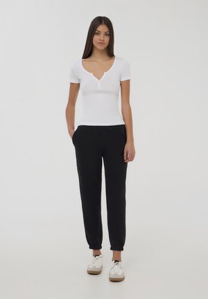 Biała bluzka z krótkim rękawem i dekoltem w serek, ozdobiona guzikami, połączona z czarnymi spodniami dresowymi z elastycznymi mankietami i kieszeniami, noszona z białymi sneakersami.