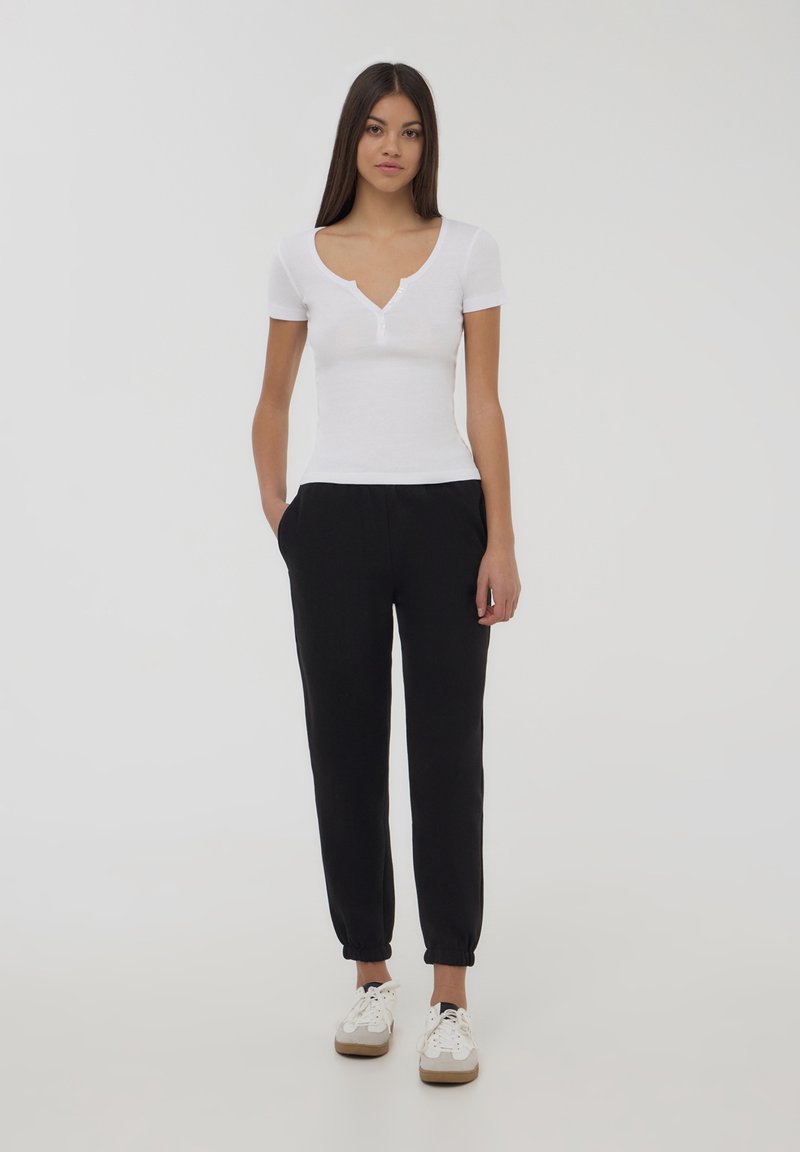 Biała bluzka z krótkim rękawem i dekoltem w serek, ozdobiona guzikami, połączona z czarnymi spodniami dresowymi z elastycznymi mankietami i kieszeniami, noszona z białymi sneakersami.