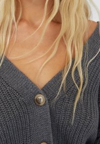 Woman wearing a dark gray knit cardigan with large brown buttons, showing a V-neckline and loose blonde hair over her shoulders.