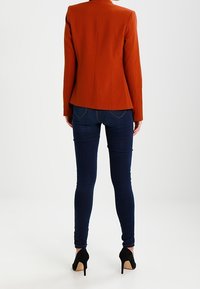Orange skräddarsydd kavaj med hög krage, figursydd design och textil med struktur, kombinerad med mörkblå slim fit-jeans och svarta högklackade skor.
