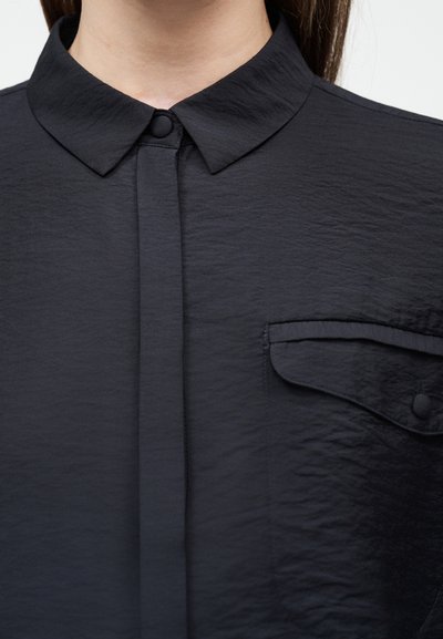 Κοντινό πλάνο μαύρου υφασμάτινου γιακά πουκάμισου με κουμπωτή λωρίδα και κουμπωτή τσέπη στο στήθος σε άτομο μπροστά σε απλό φόντο.